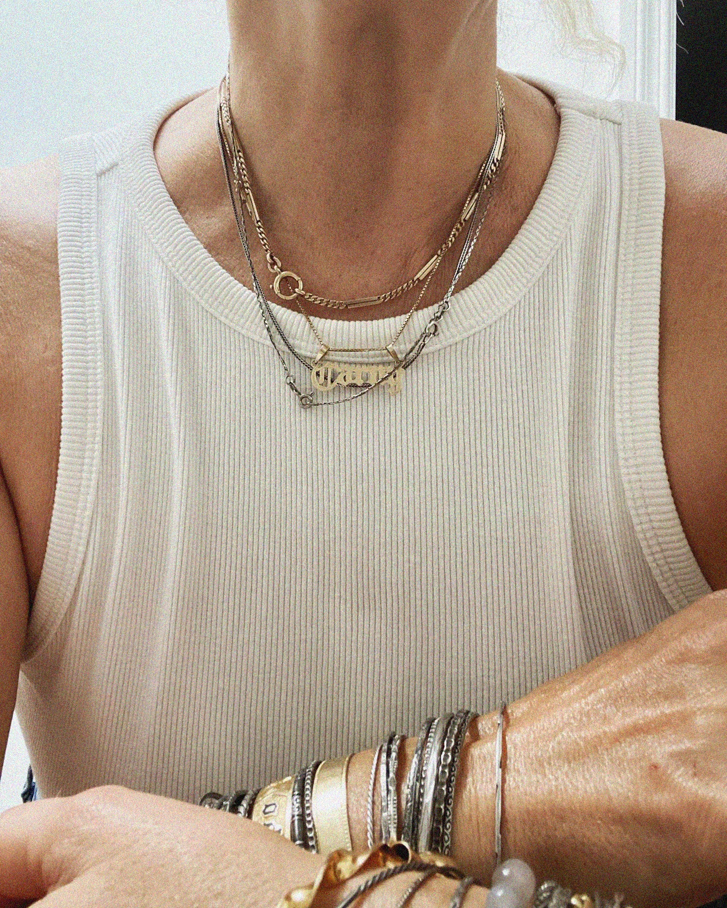 women wearing white tank top with layer gold and silver necklaces and bracelets. Arms crossed in front of body.