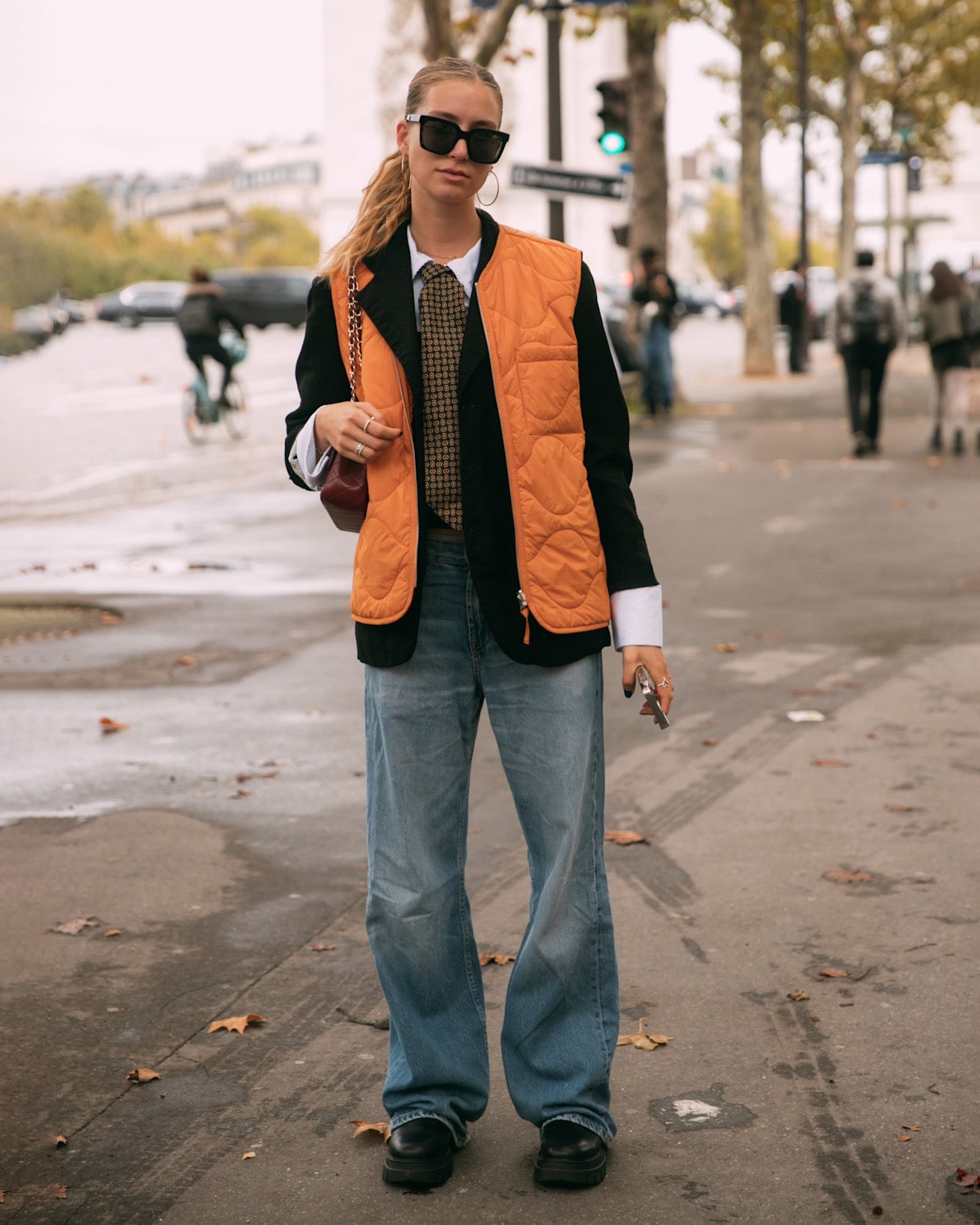 Woman in Paris wearing light wash denim with a black blazer, tie, orange vest, and chunky sunglasses.