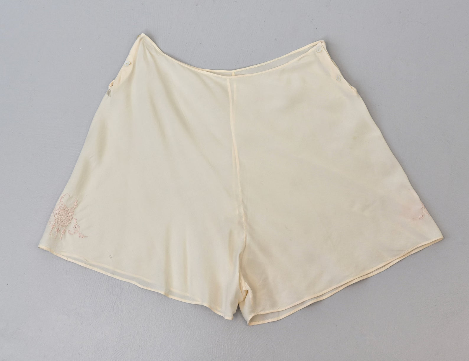  Ivory hued, in certain lighting it almost has a faint peach undertone. Pink floral embroidery detailing on each leg. Double button closures on each side of waist. Unlined